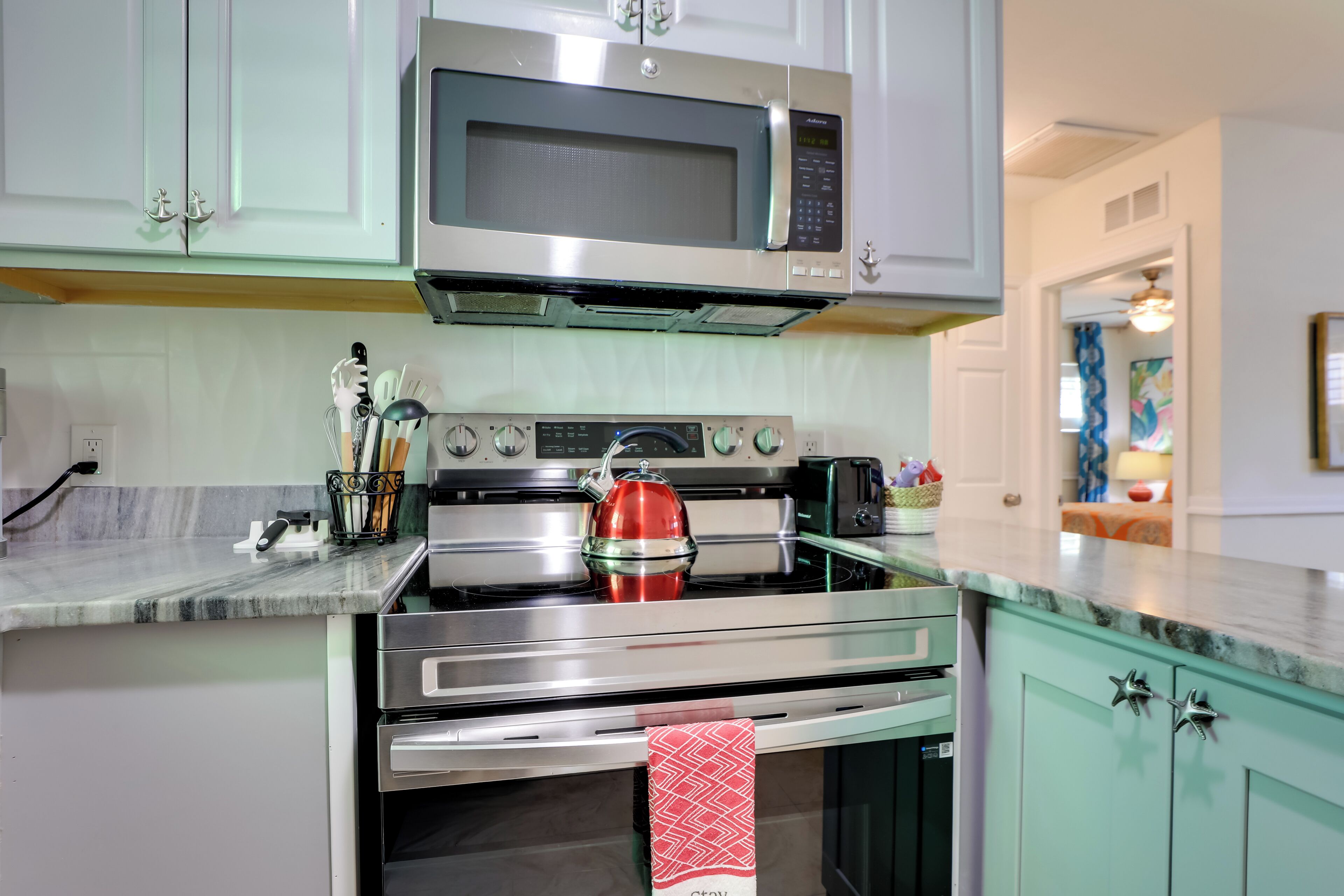 A kitchen with a stainless steel microwave, oven, and kettle. There are cabinets, a countertop, and a window.