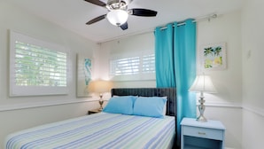 Une chambre à coucher comprenant un lit, une table de chevet, un ventilateur de plafond et une fenêtre avec des stores.