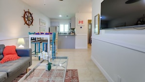 Un salon avec un canapé, une table basse et un ventilateur de plafond. On aperçoit, au fond, un coin cuisine avec une table à manger et des chaises.