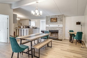 Uma cozinha espaçosa com lareira de pedra, uma sala de jantar com uma mesa comprida e cadeiras, e um lustre moderno.