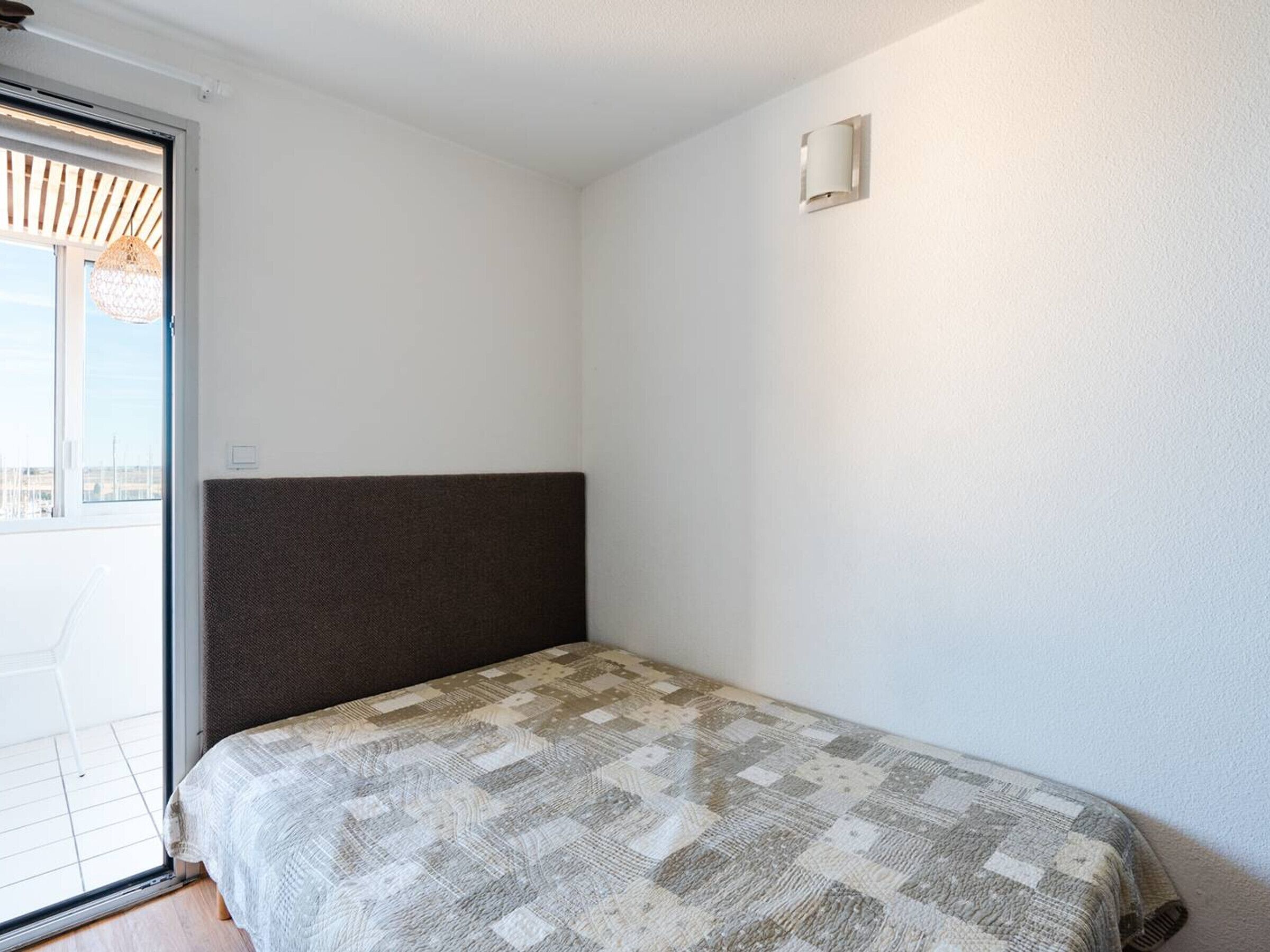Ein kleines Zimmer mit einem Einzelbett, einem dunklen Kopfteil, einer gemusterten Bettdecke und einer Glastür zum Außenbereich.