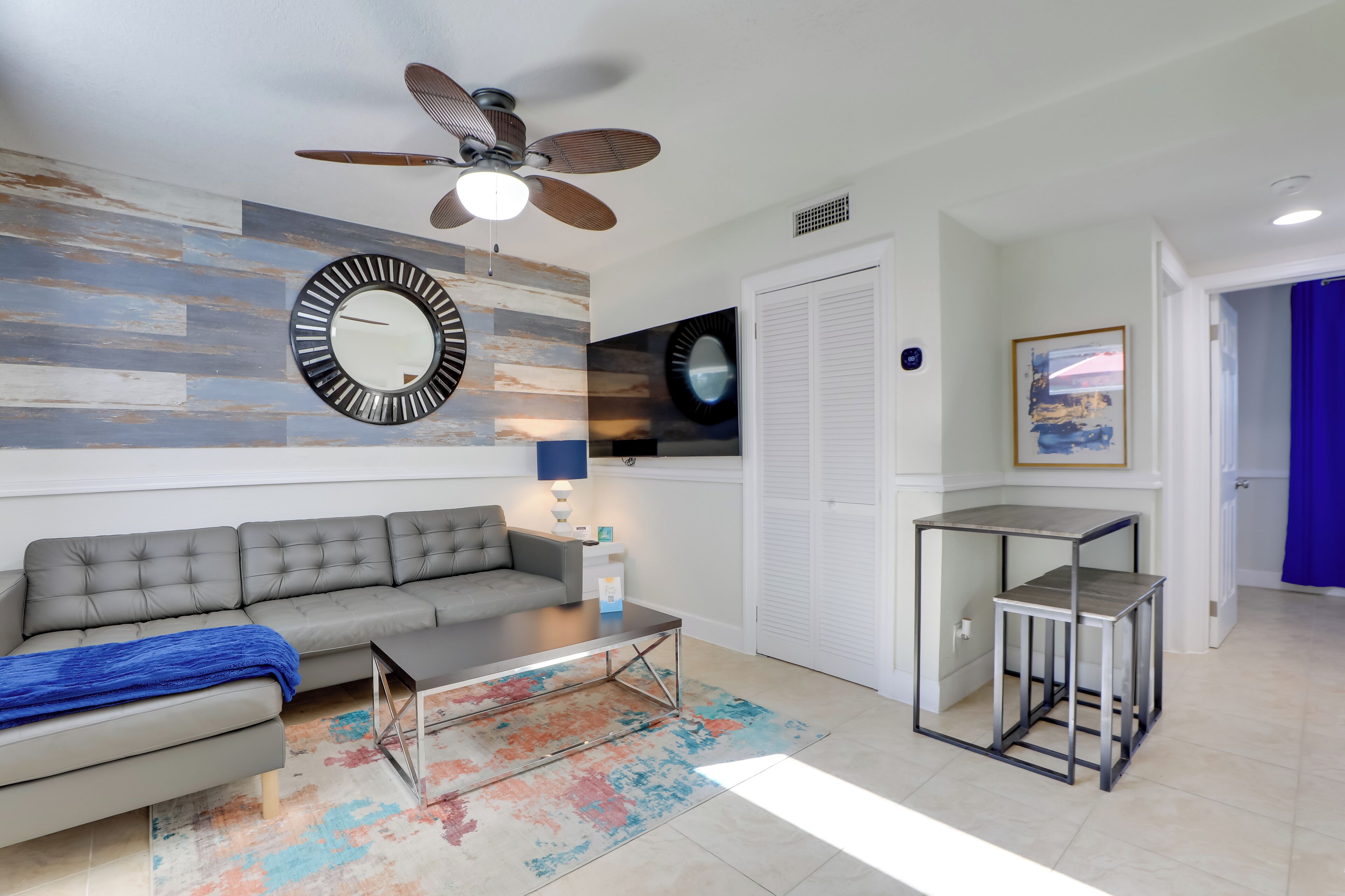 Un salon moderne avec un canapé gris, une table basse, un ventilateur de plafond et un téléviseur fixé au mur.