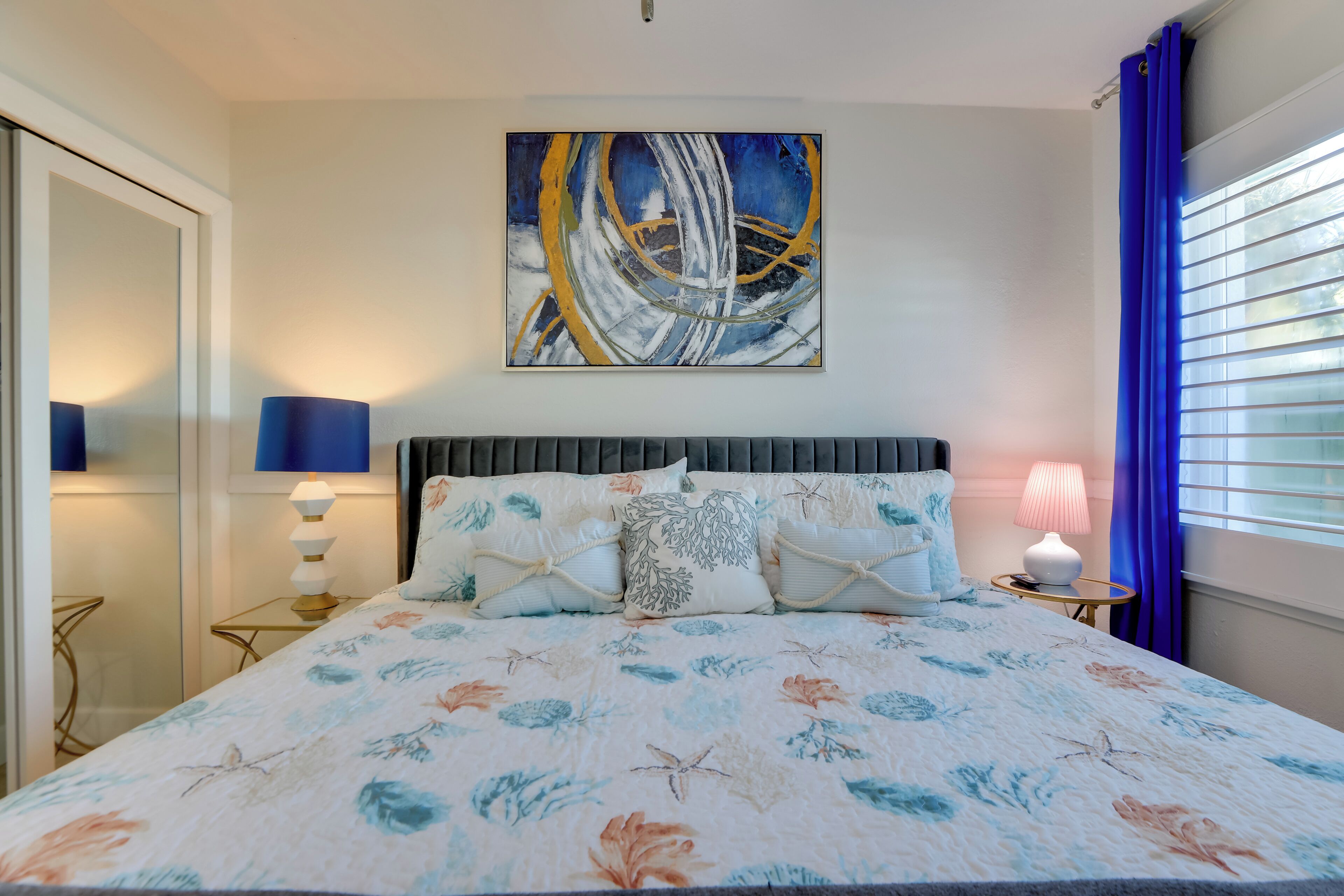 Un dormitorio con una colcha estampada floral, un cuadro abstracto grande en la pared, una mesita de noche con lámpara y una ventana con cortinas azules.