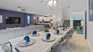 A modern dining area with a long table set for six, a kitchen in the background, and a staircase leading to another level. - Game Room, Theater & Pool! Ultimate Disney Oasis (Davenport)