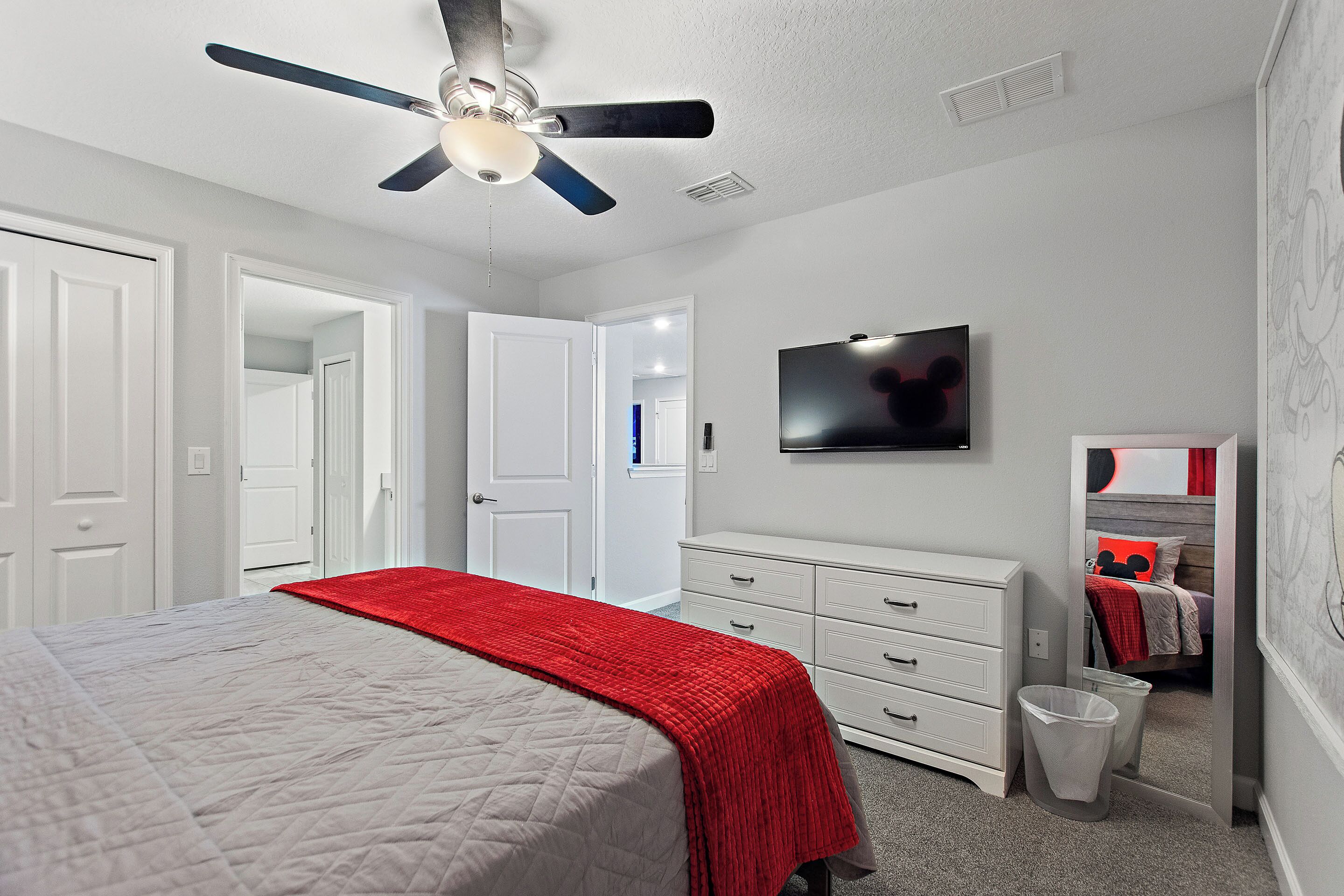 Une chambre à coucher comprenant un lit, un ventilateur de plafond, une commode, un téléviseur et une poubelle.