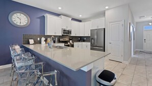 A modern kitchen with white cabinets, a stainless steel refrigerator, a microwave, and a sink. There is a large island with a sink and bar stools. - Game Room, Theater & Pool! Ultimate Disney Oasis (Davenport)
