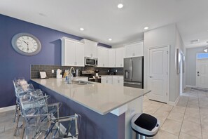 A modern kitchen with white cabinets, a stainless steel refrigerator, a microwave, and a sink. There is a large island with a sink and bar stools. - Game Room, Theater & Pool! Ultimate Disney Oasis (Davenport)