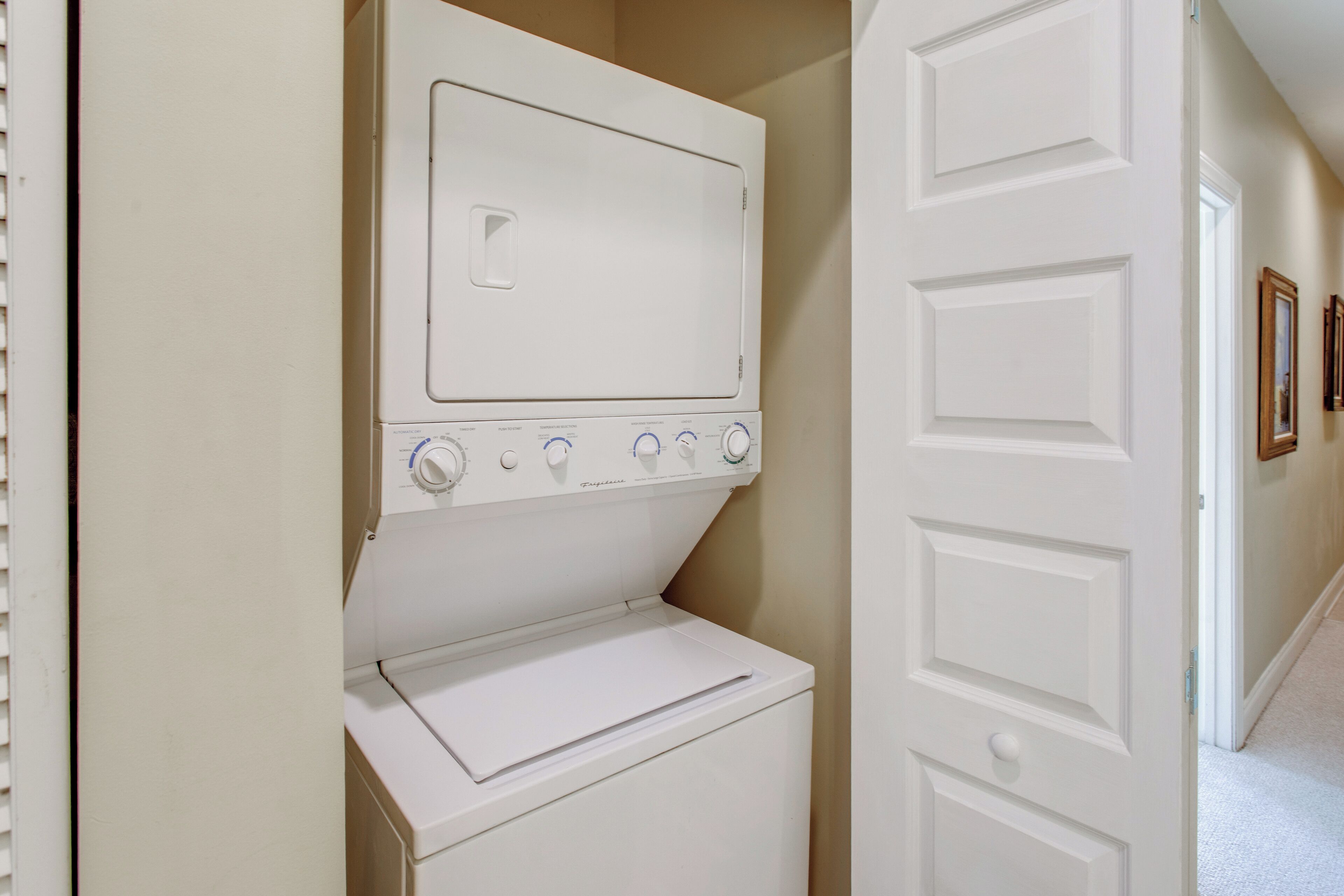 A laundry area with a front-loading washer and dryer, a white door, and a hallway with framed pictures on the wall.