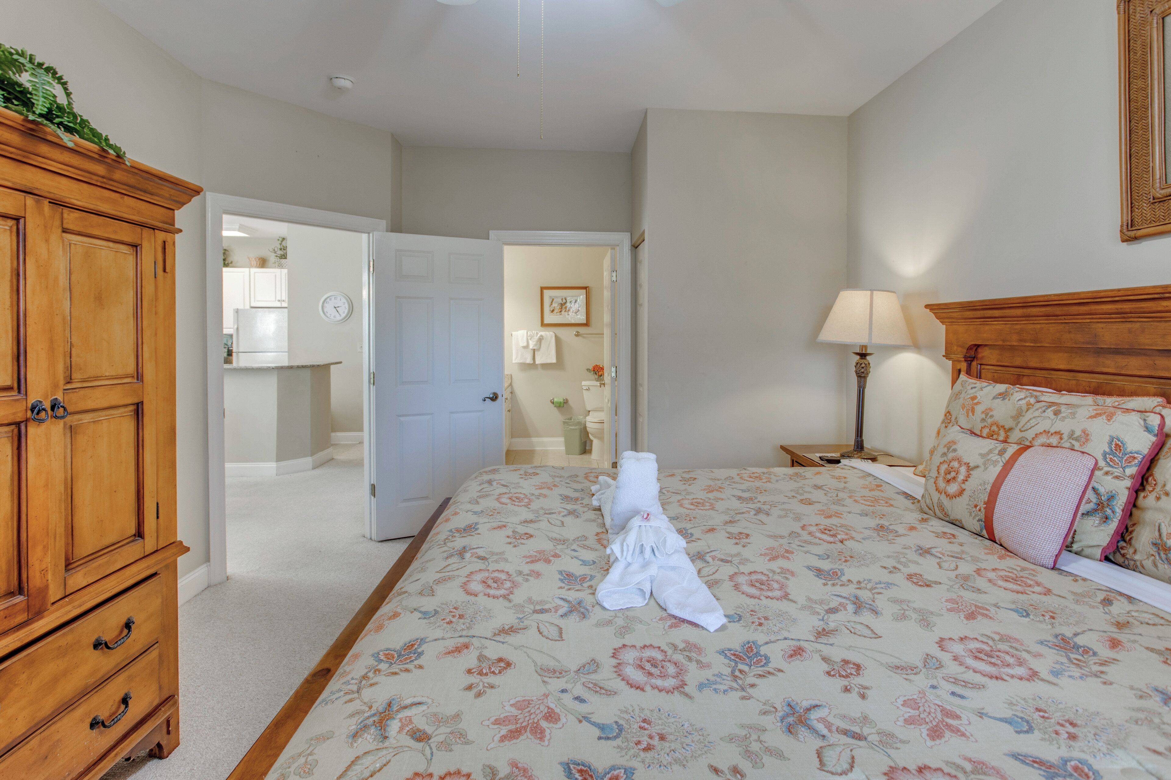 A bedroom with a floral bedspread, a wooden dresser, a nightstand with a lamp, and a door leading to a bathroom.