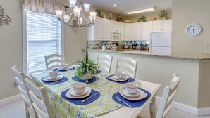 Zona pranzo con tavolo apparecchiato per quattro, sedie bianche, un lampadario e una cucina sullo sfondo.