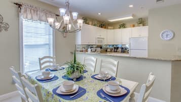 A dining area with a table set for four, white chairs, a chandelier, and a kitchen in the background.
