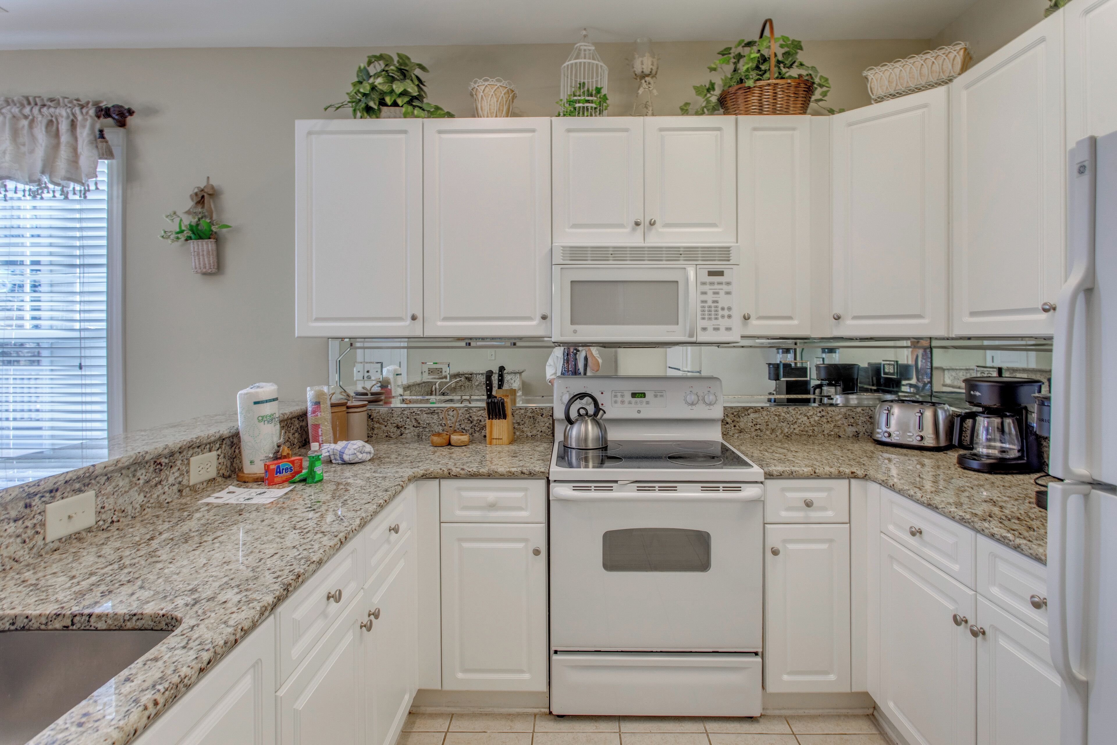 A kitchen with white cabinets, a granite countertop, a built-in microwave, and a stove. There are plants on the cabinets and a window with blinds.