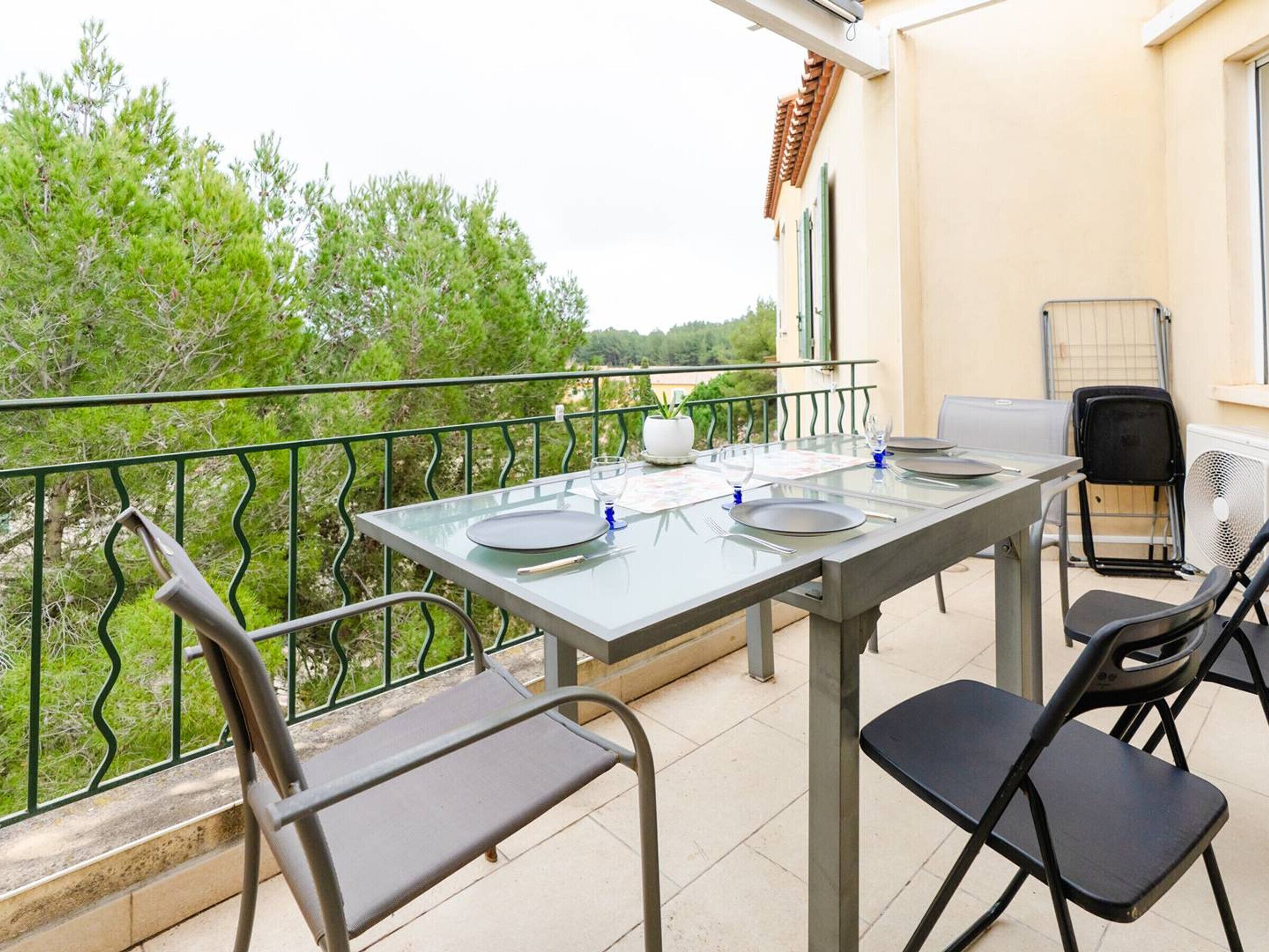 Un balcon avec une table dressée pour quatre personnes, des chaises, une plante en pot et un ventilateur.