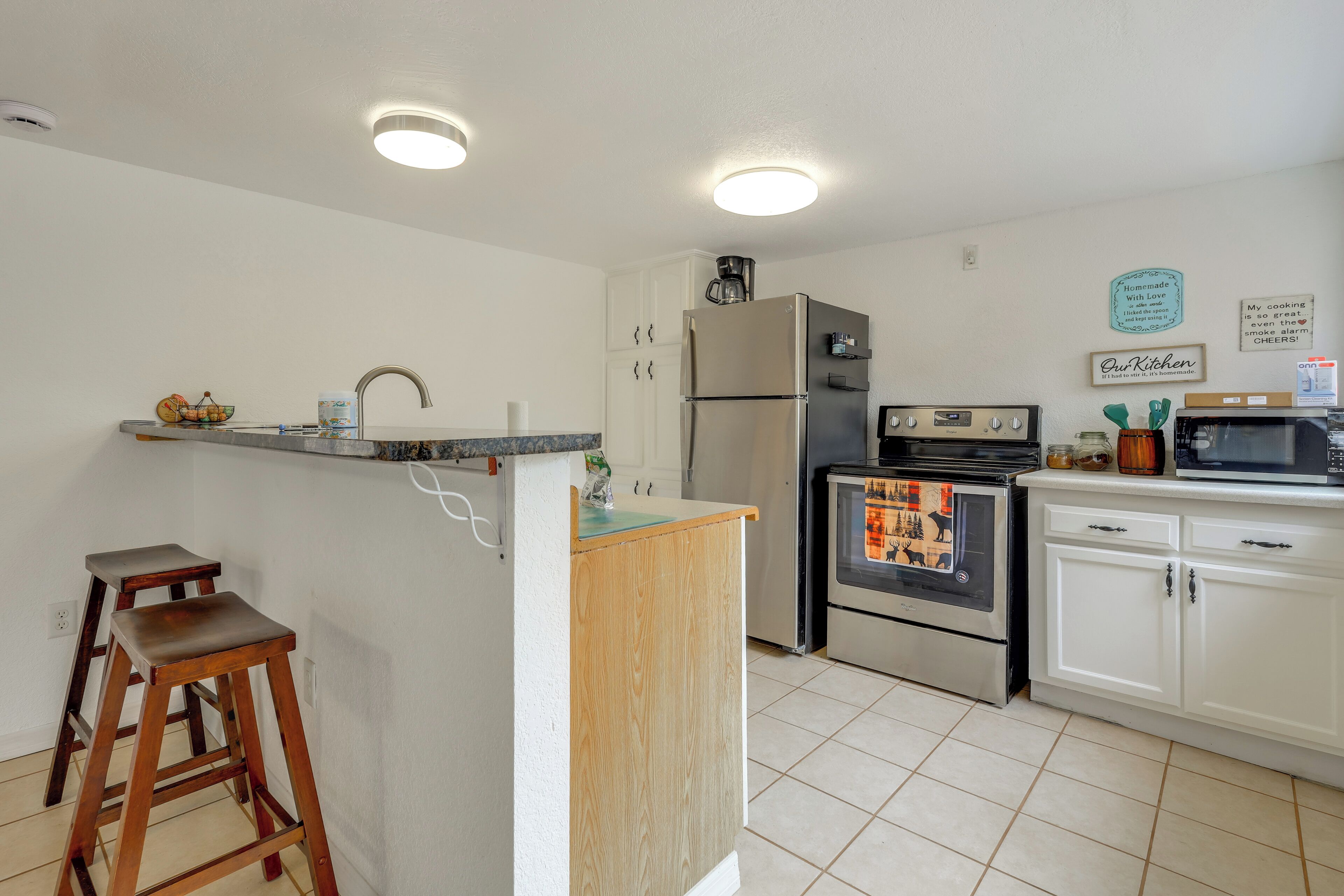Une cuisine compacte avec des meubles blancs, un réfrigérateur en acier inoxydable, un four à micro-ondes et une cuisinière. On y trouve deux tabourets en bois et un petit îlot central en bois équipé d’un évier.