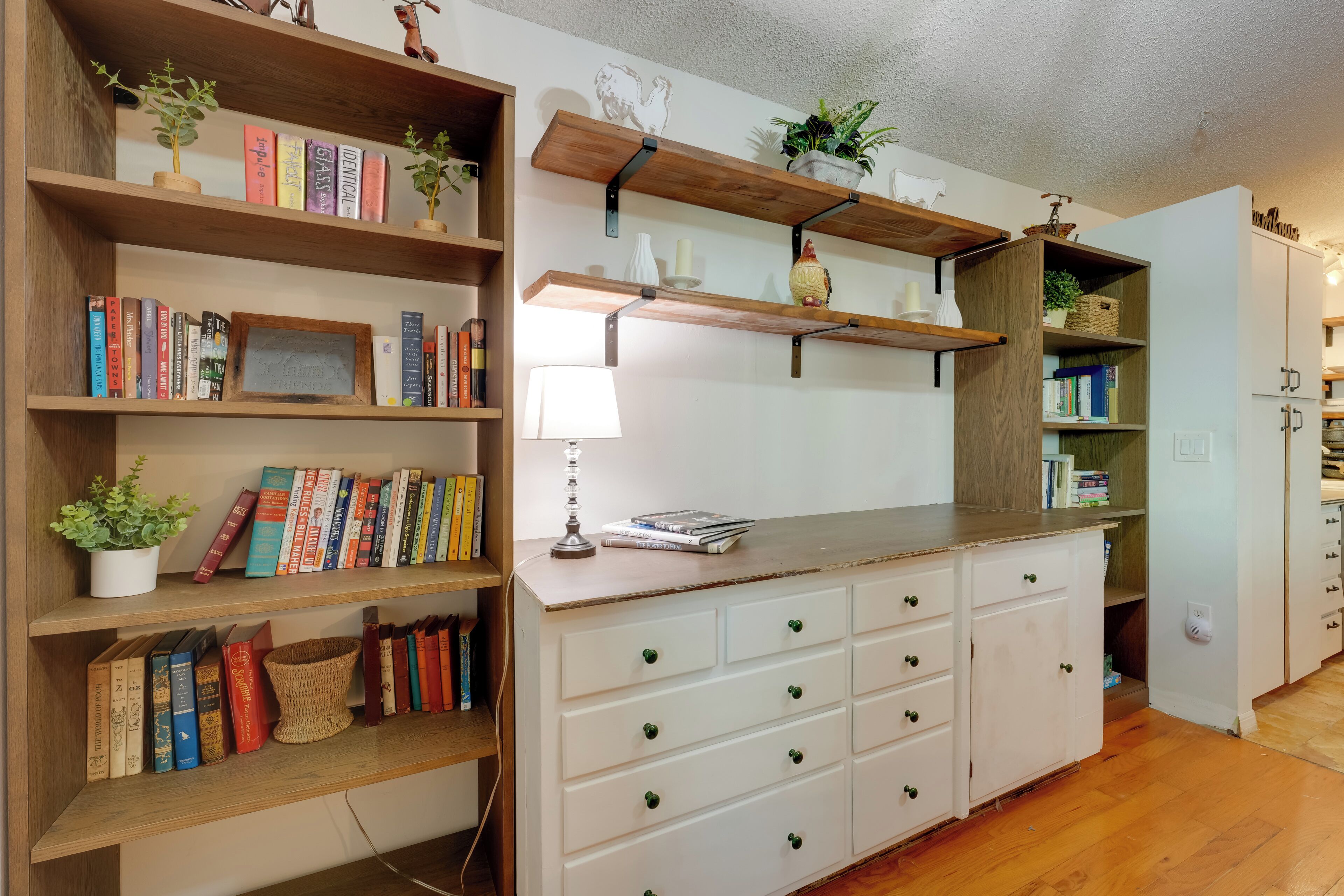 Una habitación con estanterías de madera, un escritorio blanco con cajones y una lámpara.
