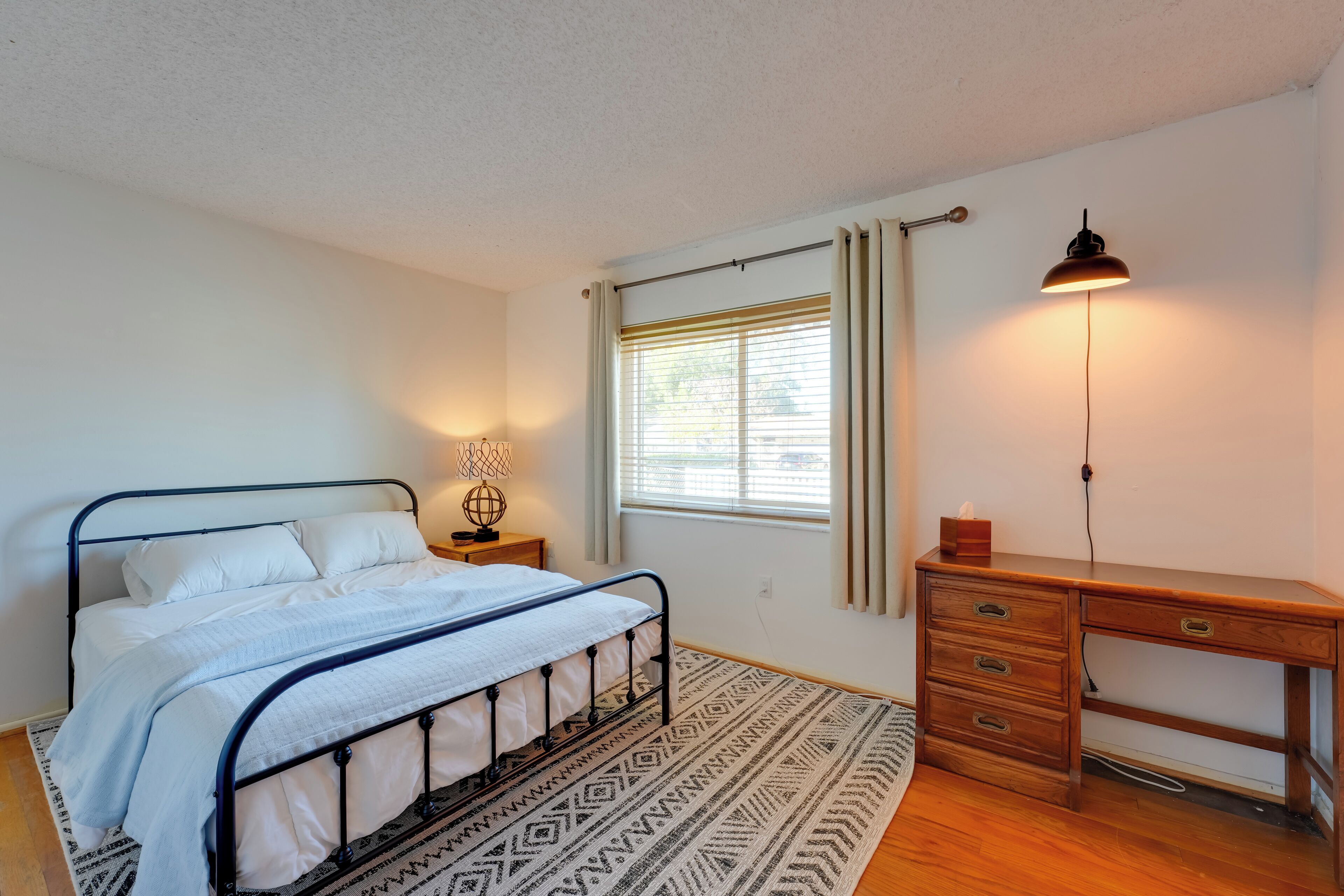 A bedroom with a bed, bedside table, lamp, window, and desk.