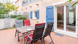 An outdoor patio with a table set for two, surrounded by chairs, and a sliding glass door leading to an interior space. - 3-room villa in Narbonne-Plage, pets allowed (Narbonne-Narbonne Plage-Narbonne Plage)