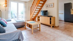 A modern living room with a wooden staircase, a TV on a wooden stand, a sofa, and a dining table. - 3-room villa in Narbonne-Plage, pets allowed (Narbonne-Narbonne Plage-Narbonne Plage)