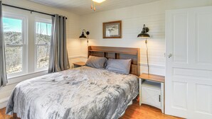 Un dormitorio con cama, dos lámparas de noche, una mesita de noche, cabecera de madera, ventana con cortinas y una puerta.