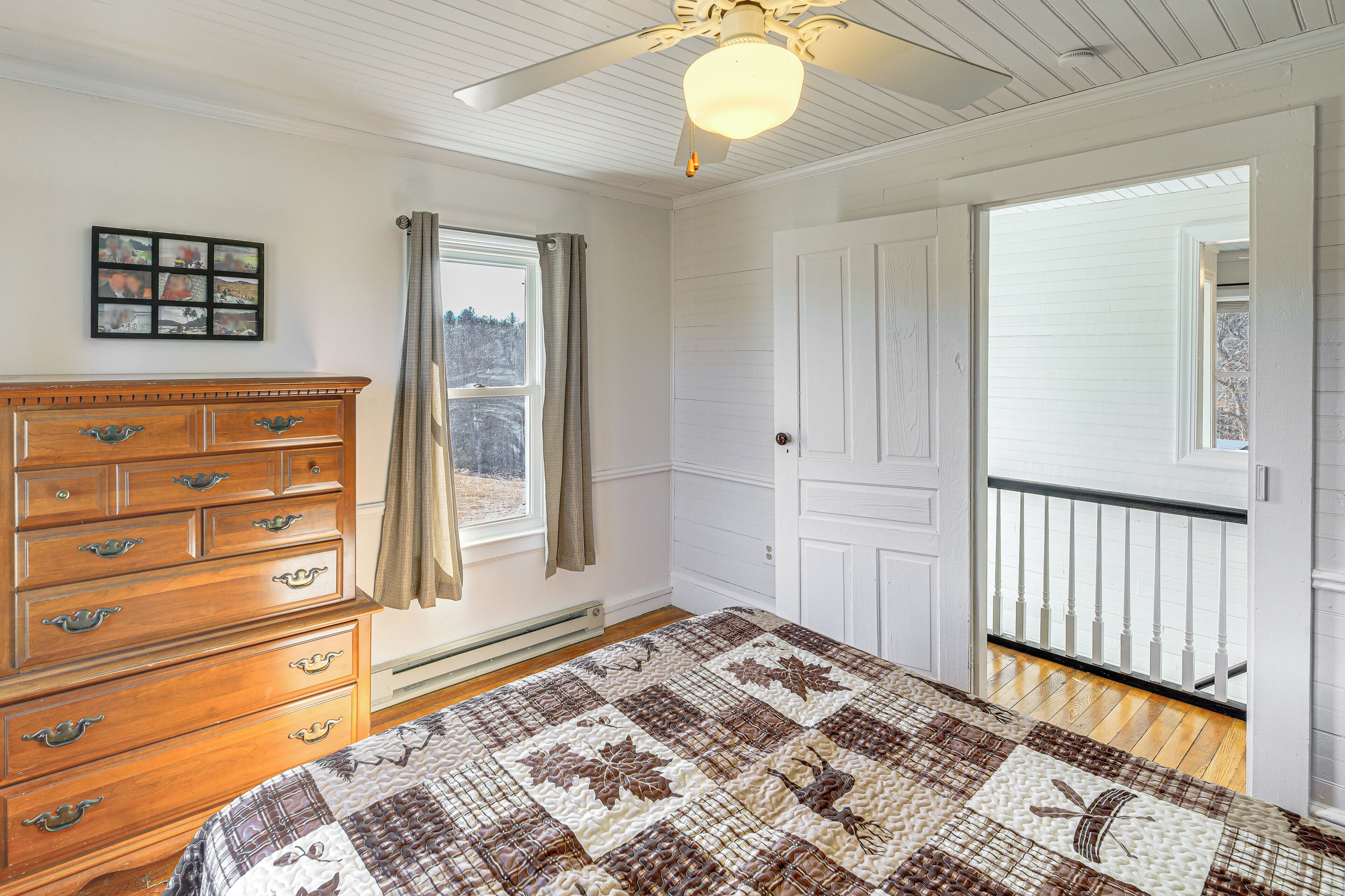 A bedroom with a wooden dresser, a quilted bedspread, a ceiling fan, and a window with curtains.