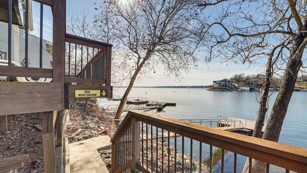 Une terrasse en bois surplombant un lac, avec un quai et des bateaux, un escalier en bois et un panneau d’avertissement indiquant de faire attention à sa tête.