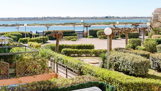 A landscaped garden with a view of the sea, featuring neatly trimmed hedges, a wooden bridge, and a pathway leading to a gate. - Seaside Apartment in Gruissan with Parking & WiFi Option (Gruissan)