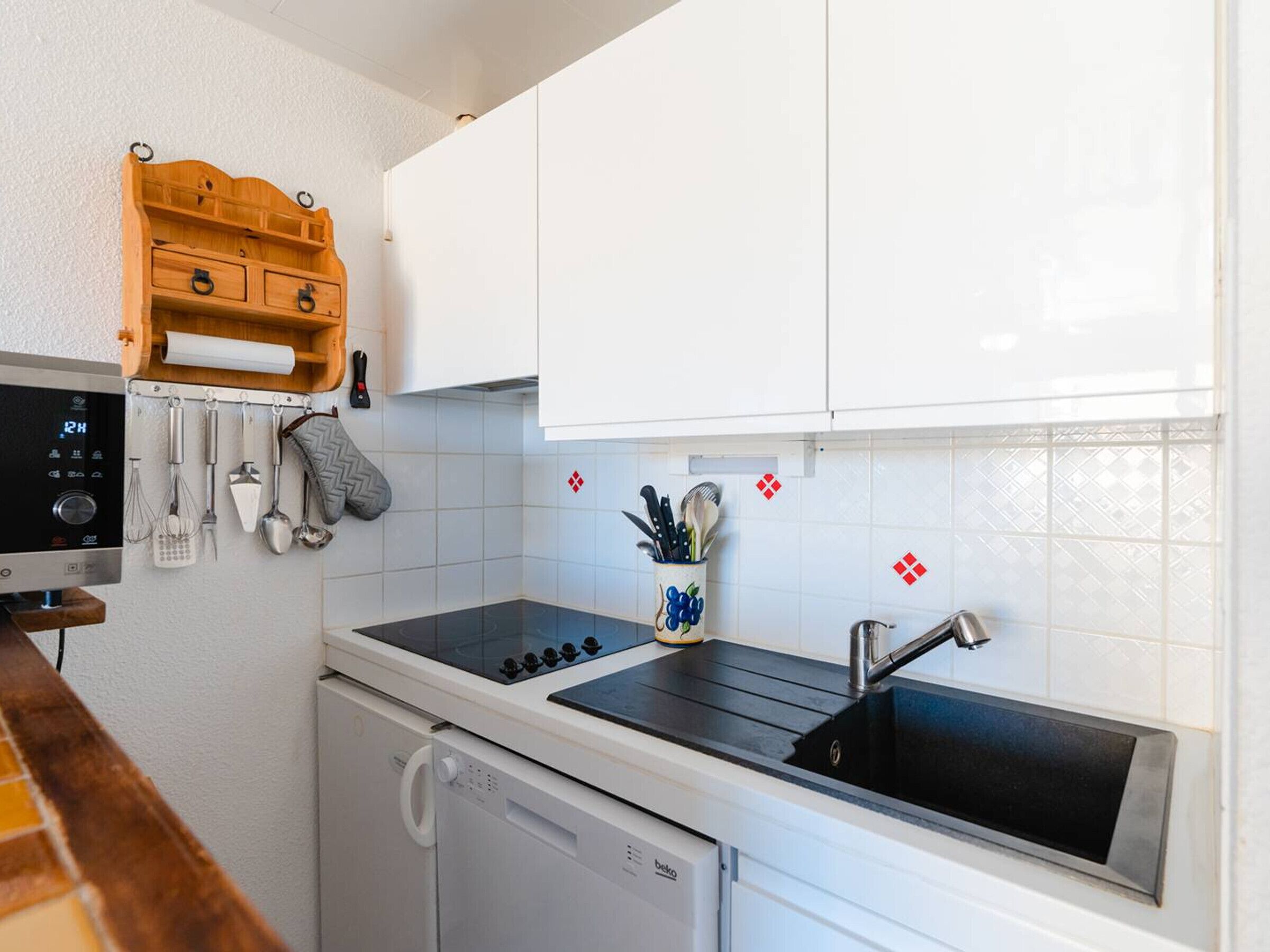 A compact kitchen with white cabinets, a black countertop, a built-in dishwasher, and a wall-mounted utensil rack.