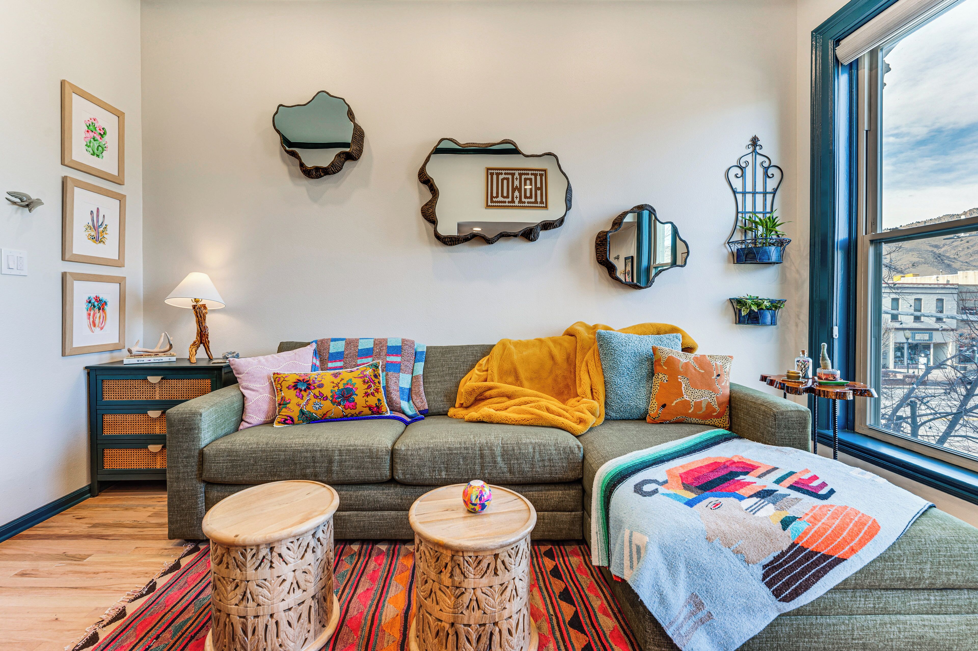 A cozy living room with a grey sofa, colorful cushions, a yellow throw blanket, and a view of a cityscape through a large window.