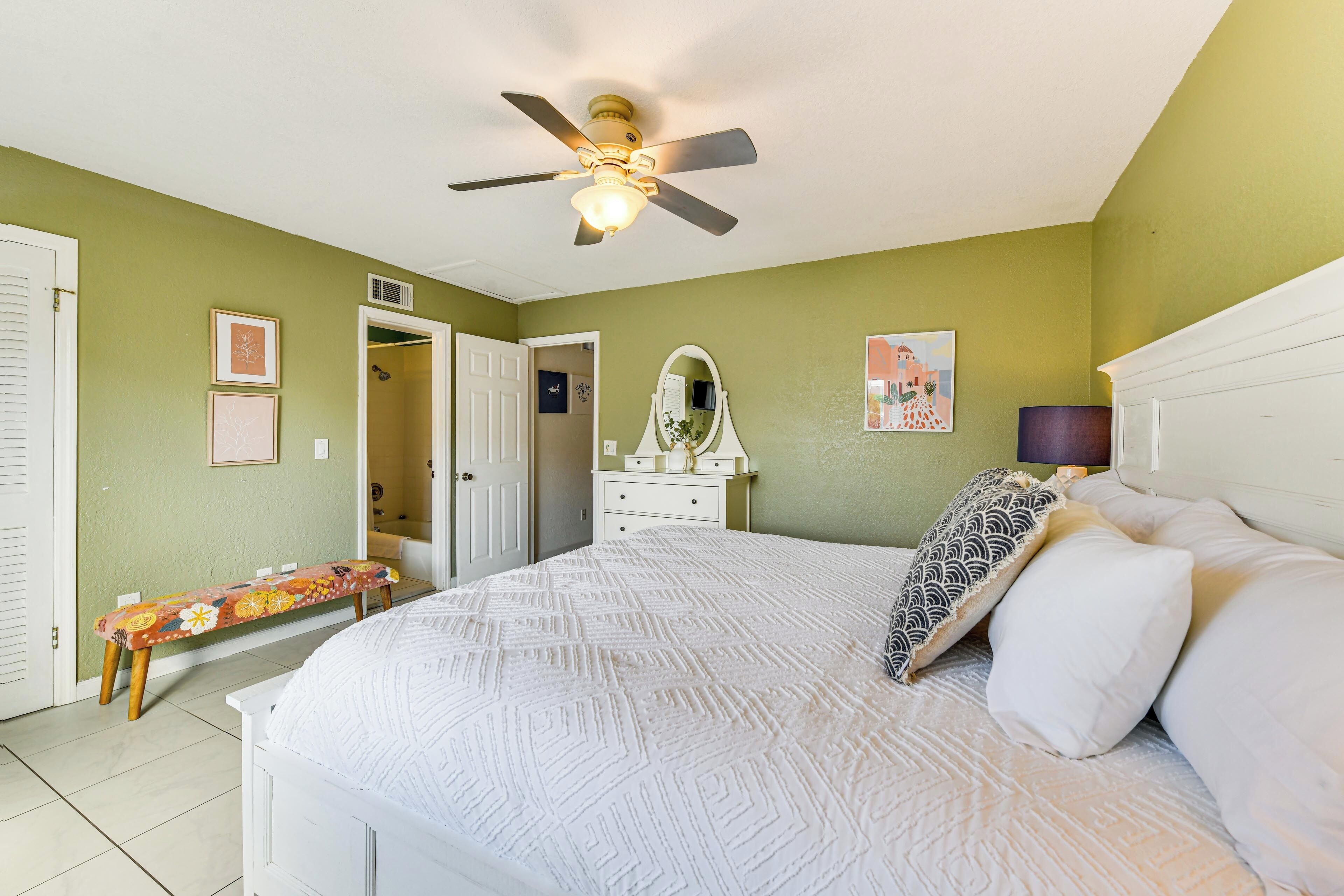 A bedroom with a bed, a ceiling fan, a bench, a dresser, and a door leading to another room.