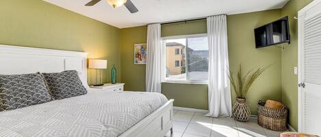 A bedroom with a bed, a ceiling fan, a TV mounted on the wall, a window with curtains, and a view of a building outside.