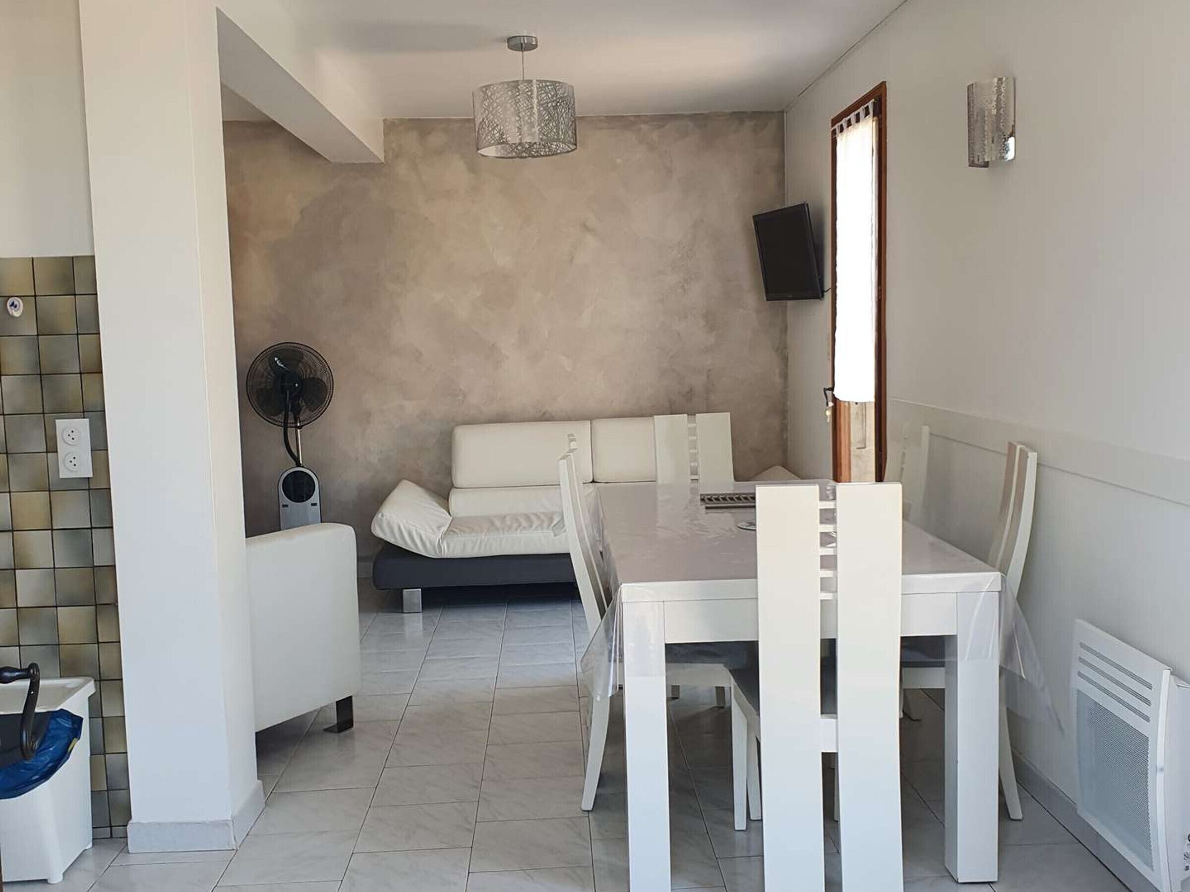 A modern living room with a white sofa, a dining table with chairs, a standing fan, and a TV mounted on the wall.