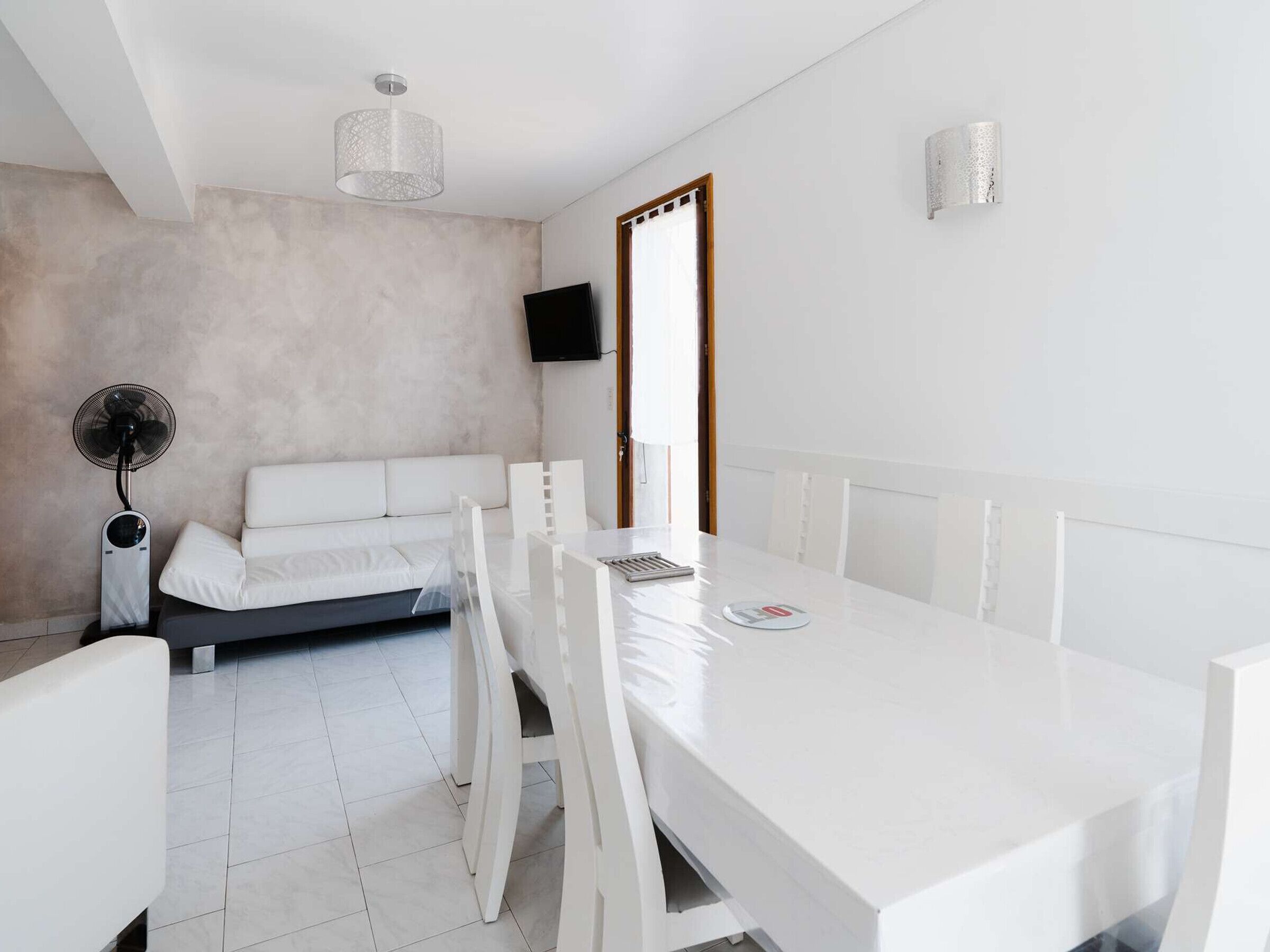 Een moderne, minimalistisch ingerichte kamer met een witte bank, eettafel en stoelen. Een plafondventilator en een aan de muur bevestigde televisie zijn zichtbaar.