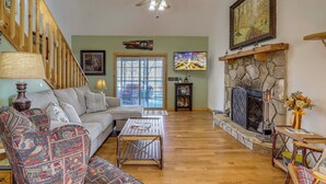A spacious living room with a wooden staircase, a stone fireplace, a large window, and a television. - Mtn-view Cabin w/ Hot Tub Near Blairsville Trails (Blairsville)