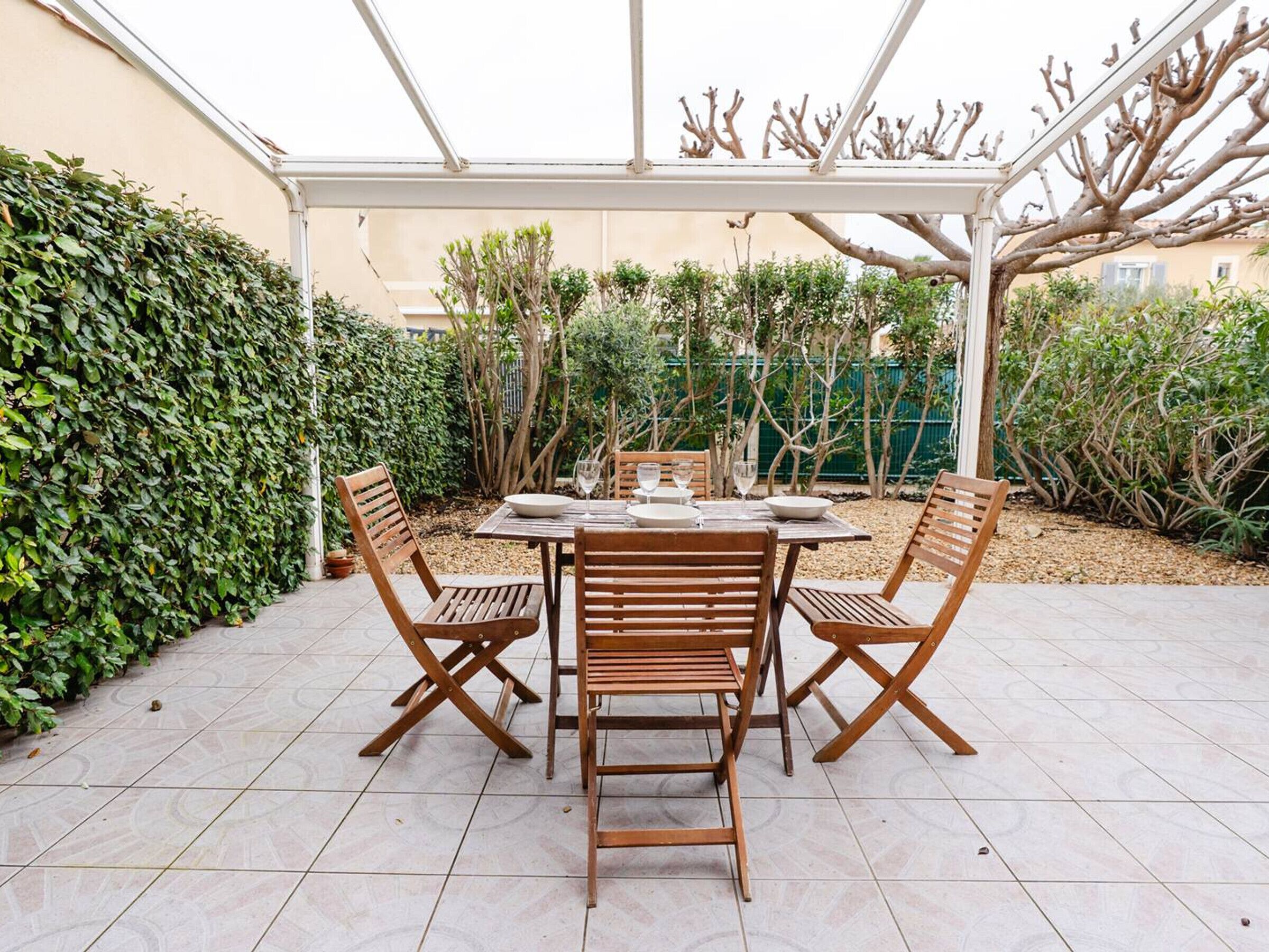 A covered patio with a table and chairs, surrounded by greenery and a tiled floor.