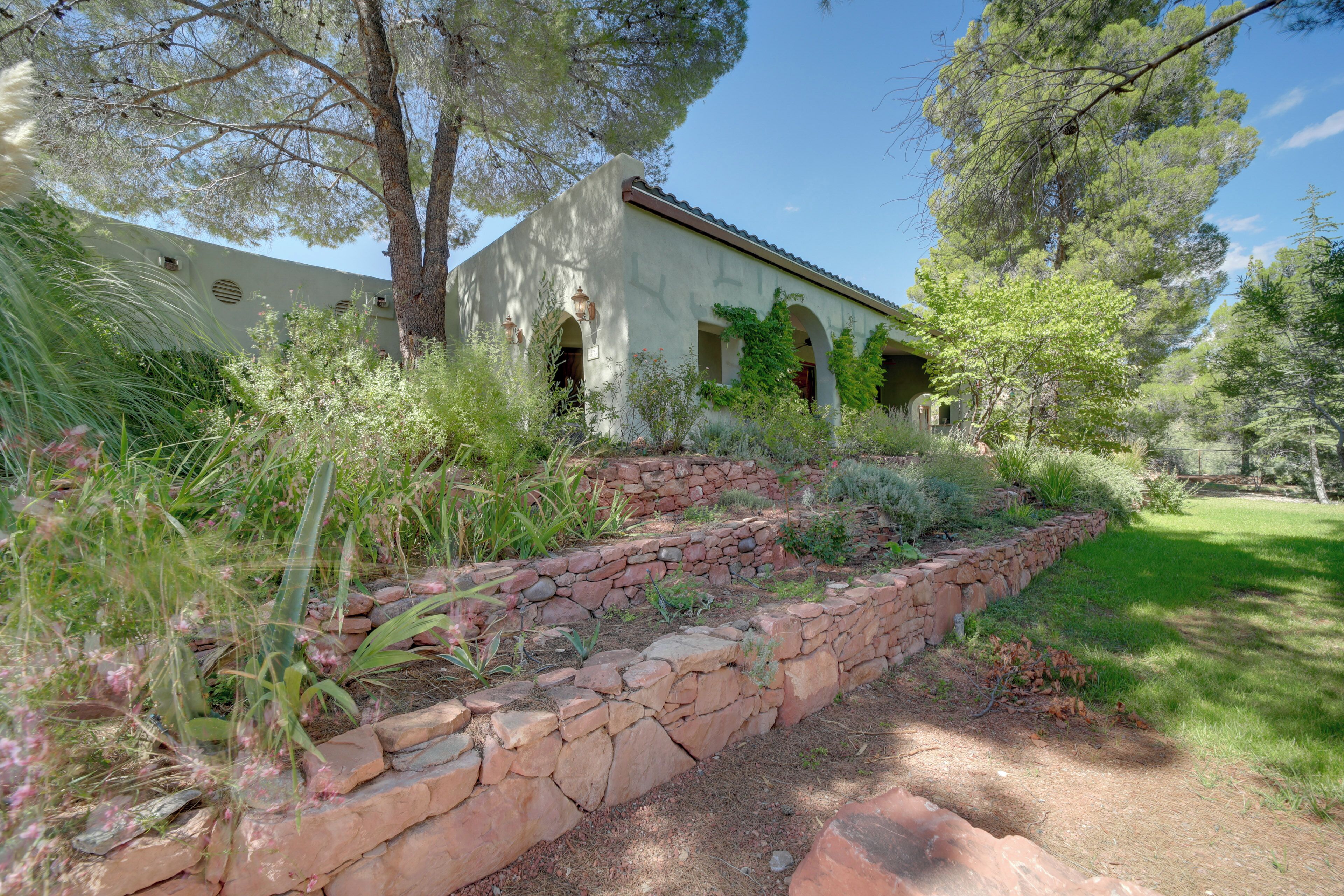 A stone retaining wall with a garden in front of a building with arches and a tiled roof.