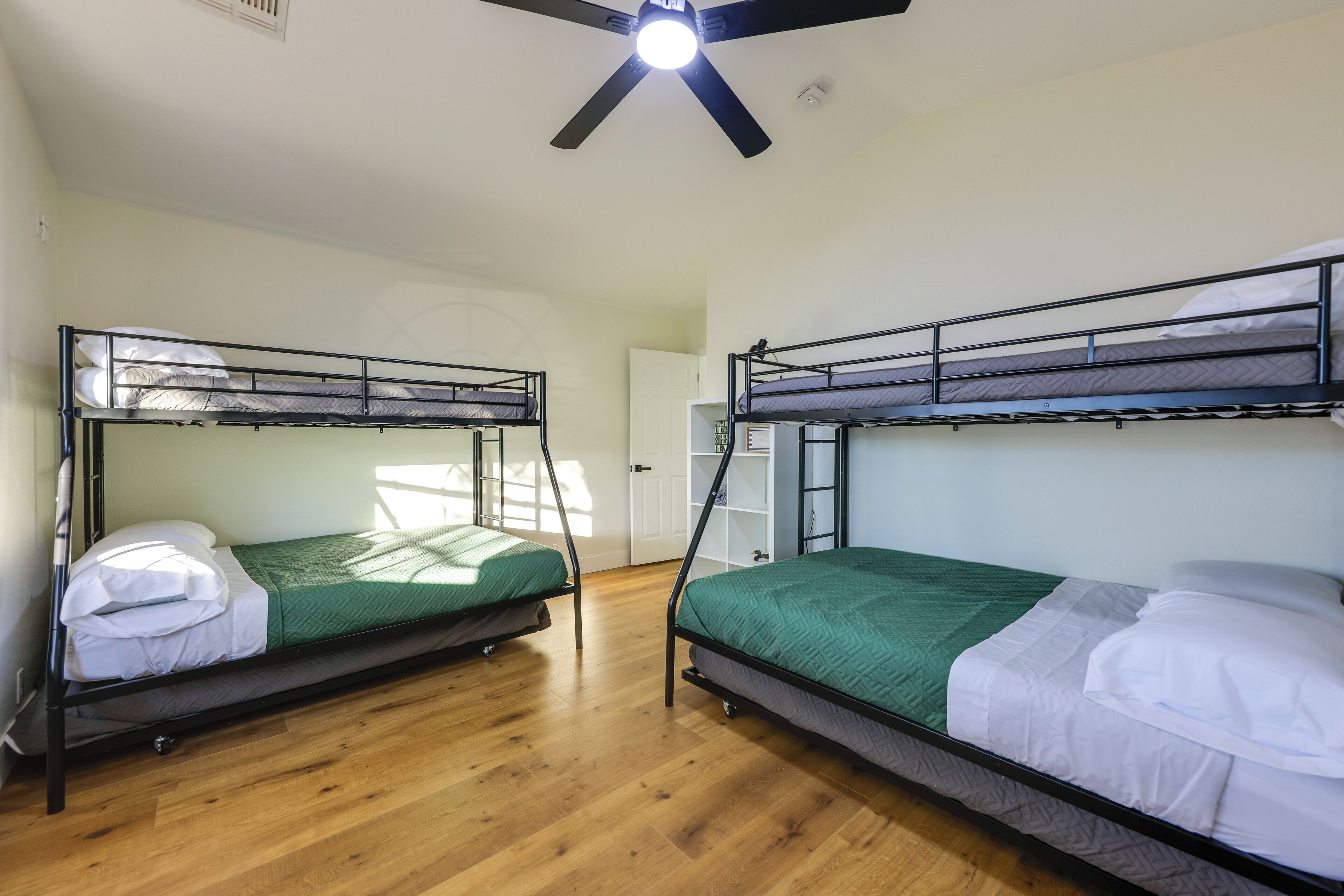A room with two bunk beds, wooden flooring, a ceiling fan, and a white door.
