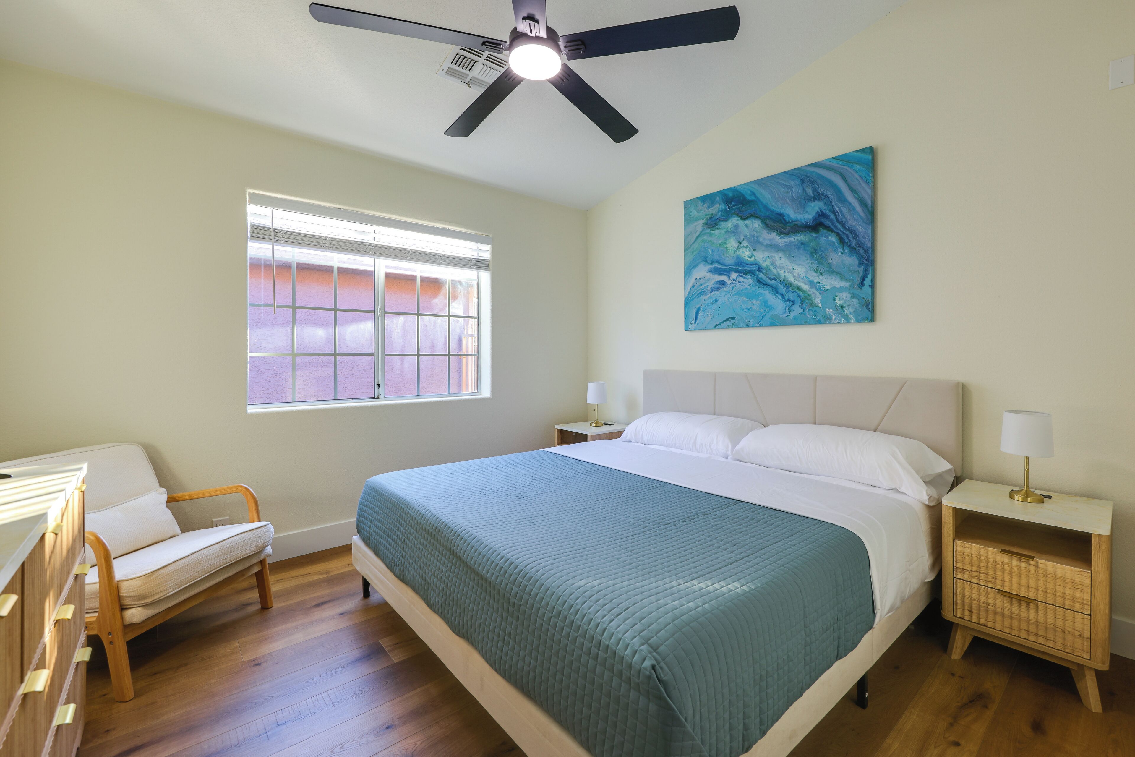 A bedroom with a bed, a nightstand, a chair, a ceiling fan, a window, and a painting on the wall.