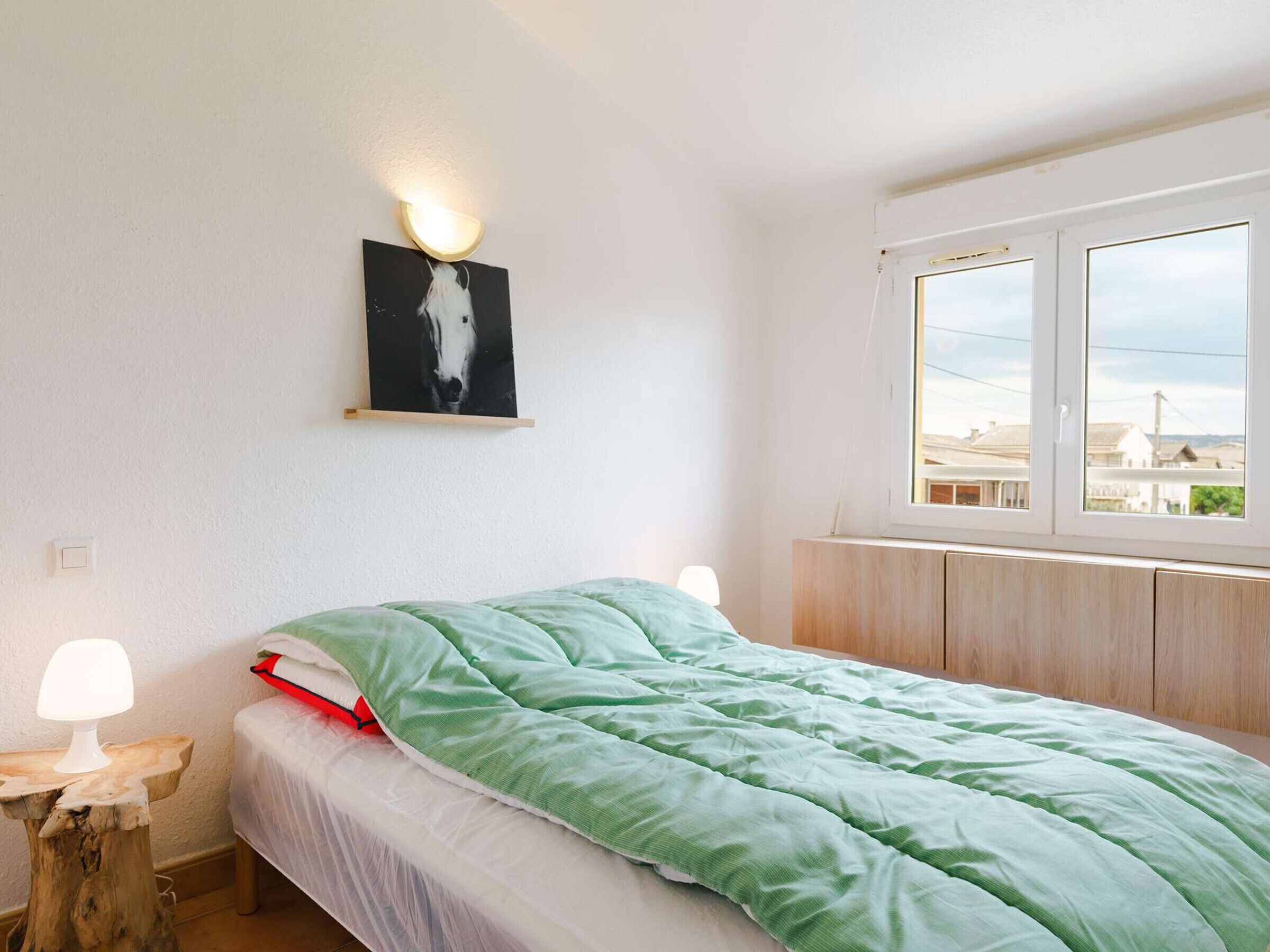 Une chambre à coucher avec un lit, une table de chevet, une lampe fixée au mur, une fenêtre donnant sur un paysage et un tableau accroché au mur.