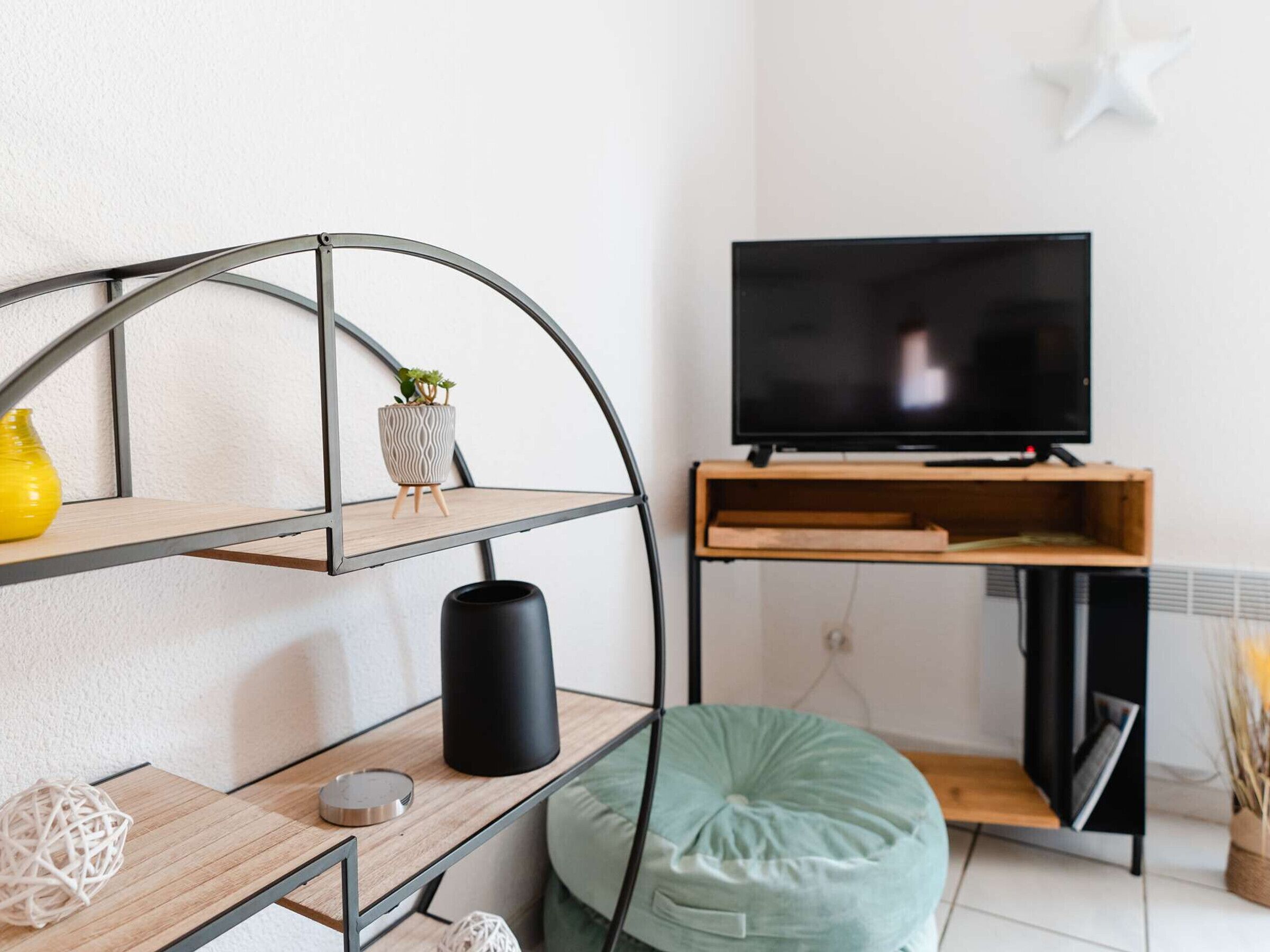 Un saló modern amb un moble prestatge metàl·lic corbat, un televisor de pantalla plana sobre un suport de fusta, un seient informal de color verd i una petita planta en test.