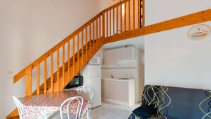 A compact living space with a wooden staircase, a dining table with chairs, a kitchenette, and a sofa. - Air-conditioned 3-Room Cottage with Mezzanine and Terrace, 200m from Narbonne Beach – Sleeps 6 (Narbonne-Narbonne Plage-Narbonne Plage)