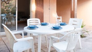 A round white table with blue plates and white plastic chairs, set on a patio with a sliding glass door leading to an indoor area. - Air-conditioned 3-Room Cottage with Mezzanine and Terrace, 200m from Narbonne Beach – Sleeps 6 (Narbonne-Narbonne Plage-Narbonne Plage)