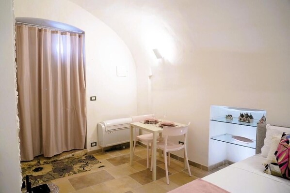 A small, well-lit room with a dining table and chairs, a bed with a colorful pillow, and a glass display case with figurines.