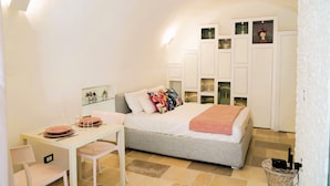 A bedroom with a bed, a small table with chairs, a wardrobe, and a wall-mounted shelf with decorative items. - La Chianca Rosa - Arcuève (Ostuni)