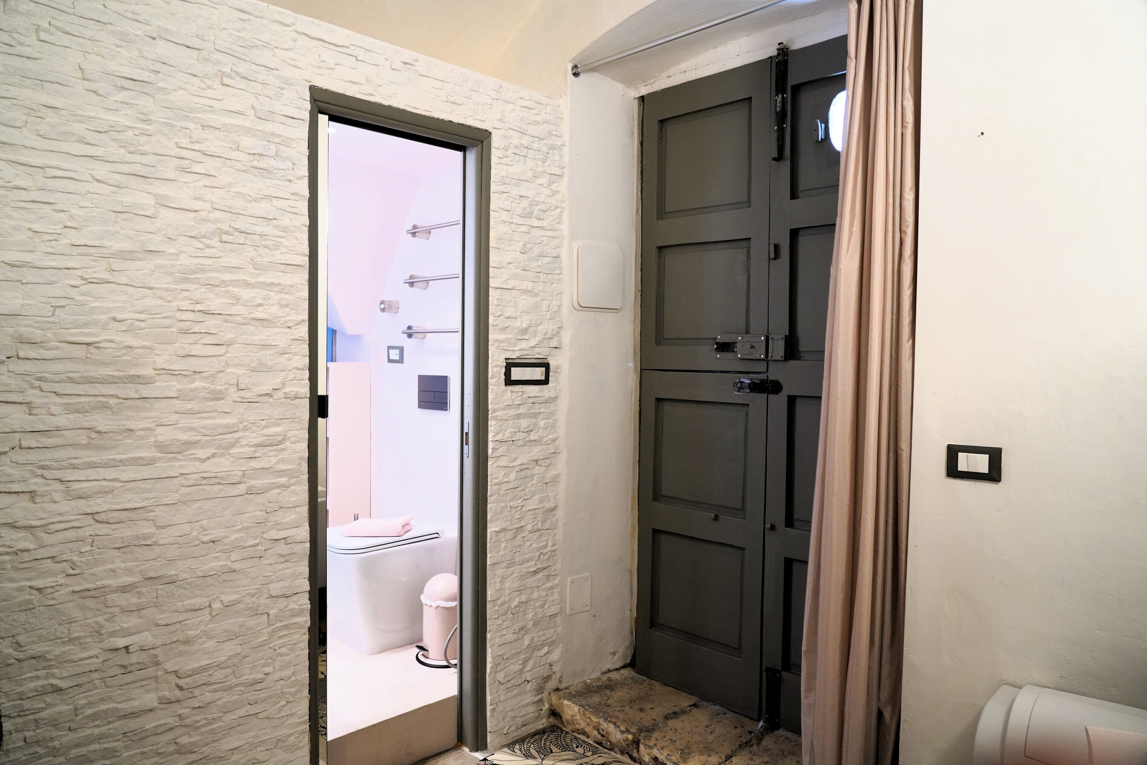 Un pasillo con pared blanca texturizada, una puerta con interruptor de luz y un escalón que lleva a un cuarto con inodoro y un cesto de basura.