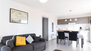 Un salon moderne avec un canapé gris, une table basse et un coin repas, la cuisine étant visible au fond.