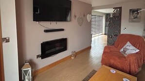 Una sala de estar con una televisión de pantalla plana montada en la pared, una chimenea debajo y un sillón naranja acogedor con un cojín.