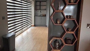 Un interior moderno con una estantería hexagonal de madera adosada a la pared, una zona de cocina con armarios grises y un ventanal con persianas horizontales.