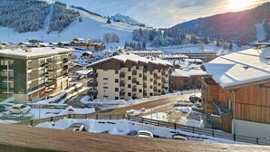 Uma cidade coberta de neve nas montanhas, com edifícios, incluindo um hotel com vários andares e varandas, e uma estação de esqui ao fundo.