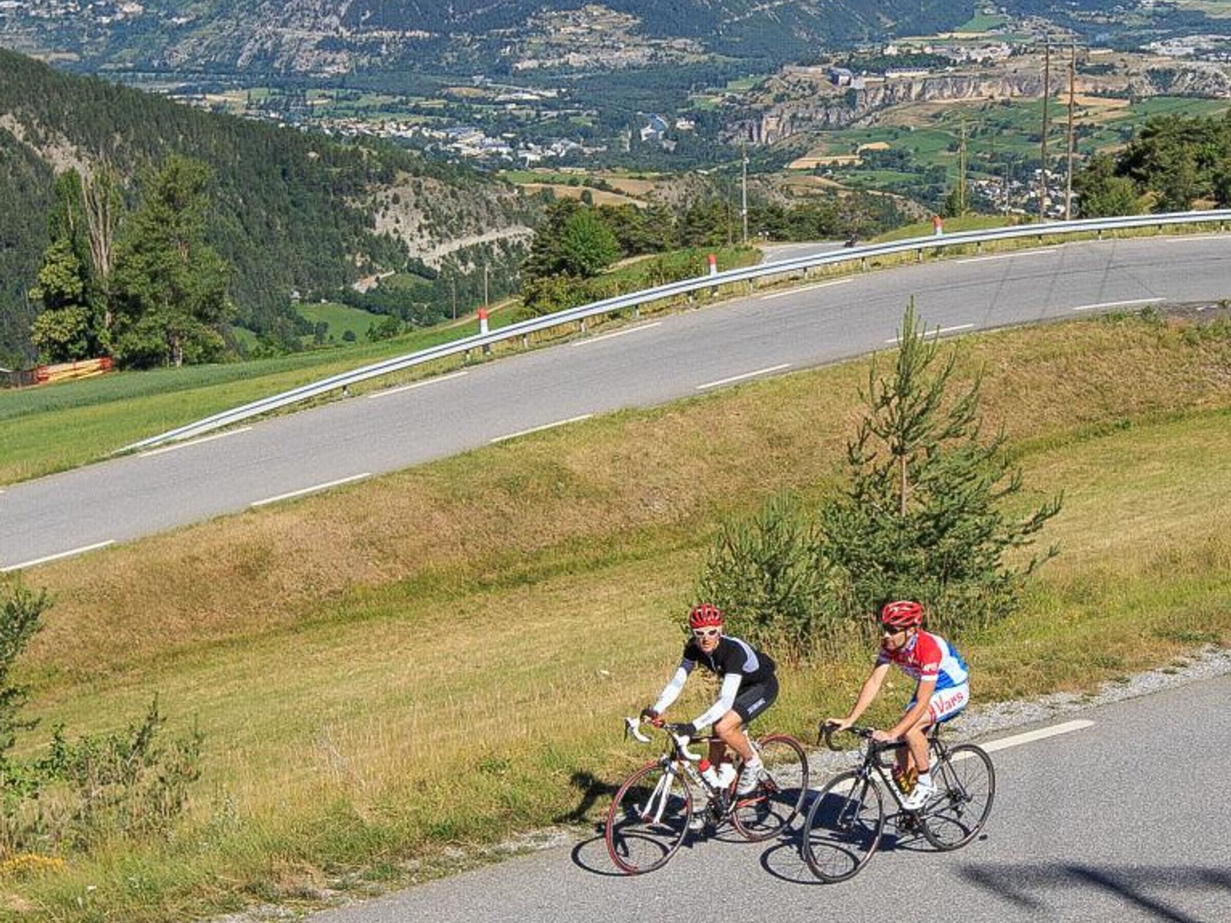 Two cyclists on a road with a guardrail, surrounded by greenery and a hilly landscape.
