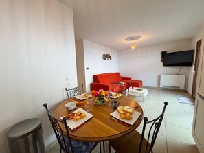 Un coin repas avec une table ronde en bois, dressée pour deux personnes, un canapé rouge et un téléviseur à écran plat fixé au mur.